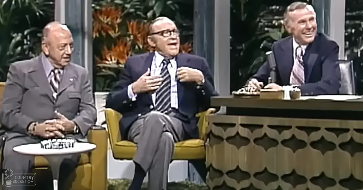 A daytime talk show set with three older adult male hosts seated in yellow armchairs and a dark wooden desk with a microphone, surrounded by greenery and bright lighting typical of mid-20th century television production.