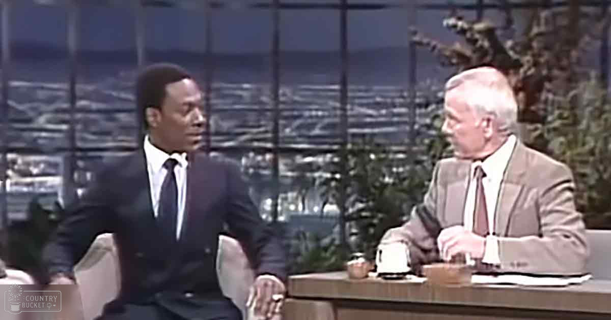 A television studio setting with two adults seated at a desk, one in a dark suit and white shirt, appearing formal, while the other wears a light-colored suit jacket over a white shirt, suggesting an older era. The backdrop shows large windows revealing a cityscape at night, indicating it's likely winter.