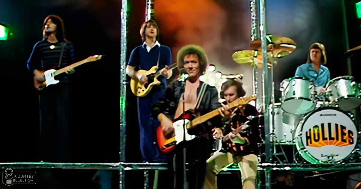 A musical performance of The Hollies at a concert venue with performers wearing dark and light-toned clothing in front of stage equipment under dim, spotlit lighting.