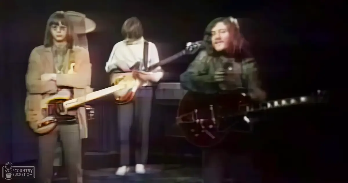 Musical performance scene from the 1960s or early 1970s, with young adult males in suits and casual shoes on stage with a dark amplifier behind them.