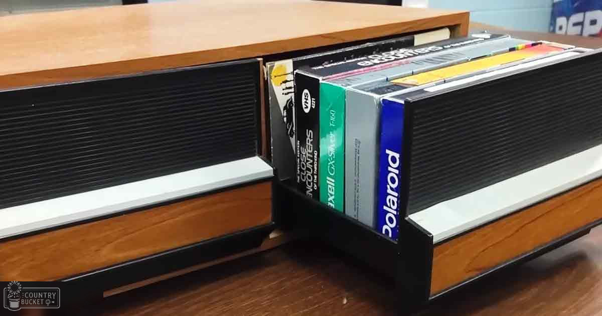 Retro-style VHS player with various colored tapes in an indoor setting, natural daylight, wooden table or shelf visible.