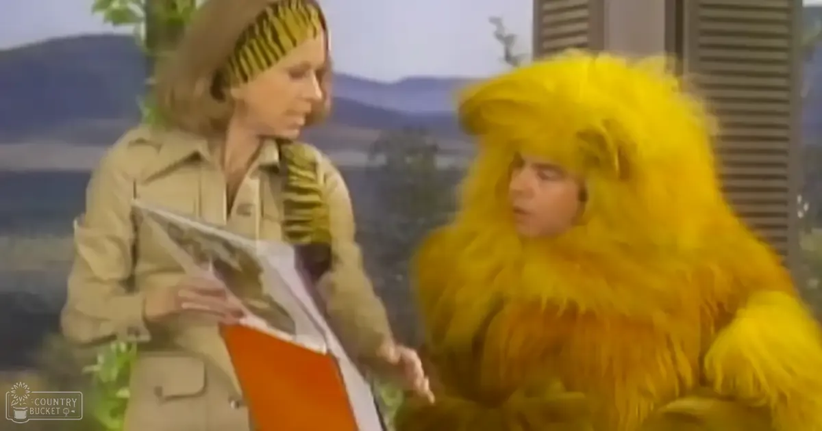 An adult female character in a tan safari-style jacket holds a book or magazine, interacting with an adult male in a large yellow lion costume against a scenic outdoor backdrop of mountains, trees, and water.