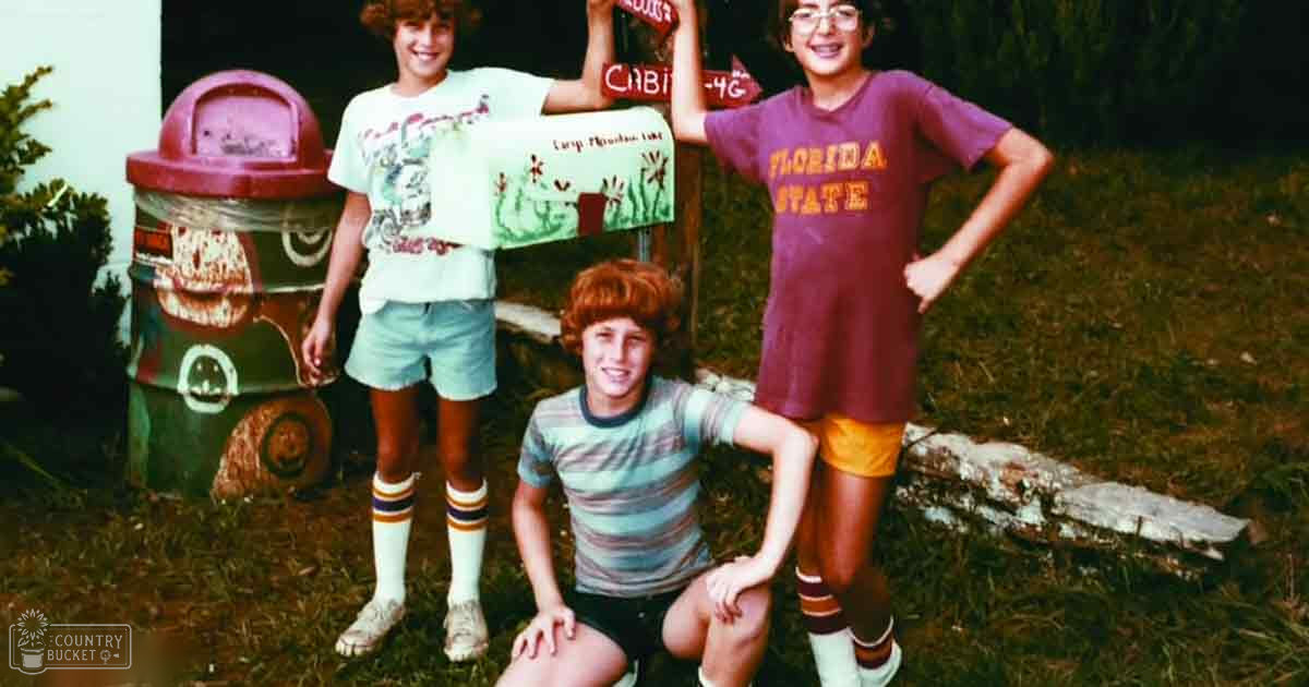 Group of children in a playful outdoor setting with creatively decorated barrel resembling a car, mailbox with drawings, grassy area. Spring or summer vibe.