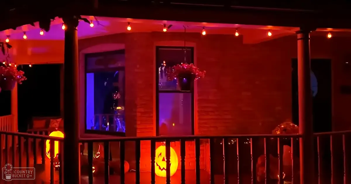 Halloween decorations on a porch at night with red lighting, potted plants, jack-o'-lanterns, and string lights creating a spooky atmosphere.