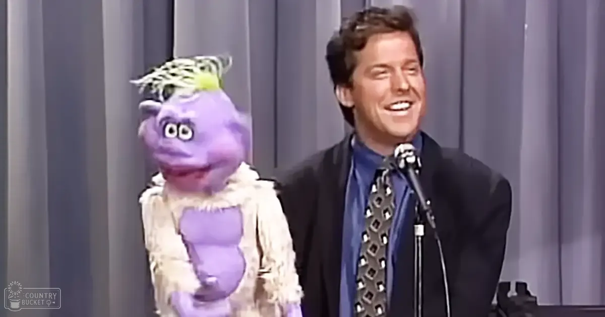 A puppet show scene with a male adult in a dark suit and geometric-patterned tie, facing a purple puppet with white fur-like texture on stage.