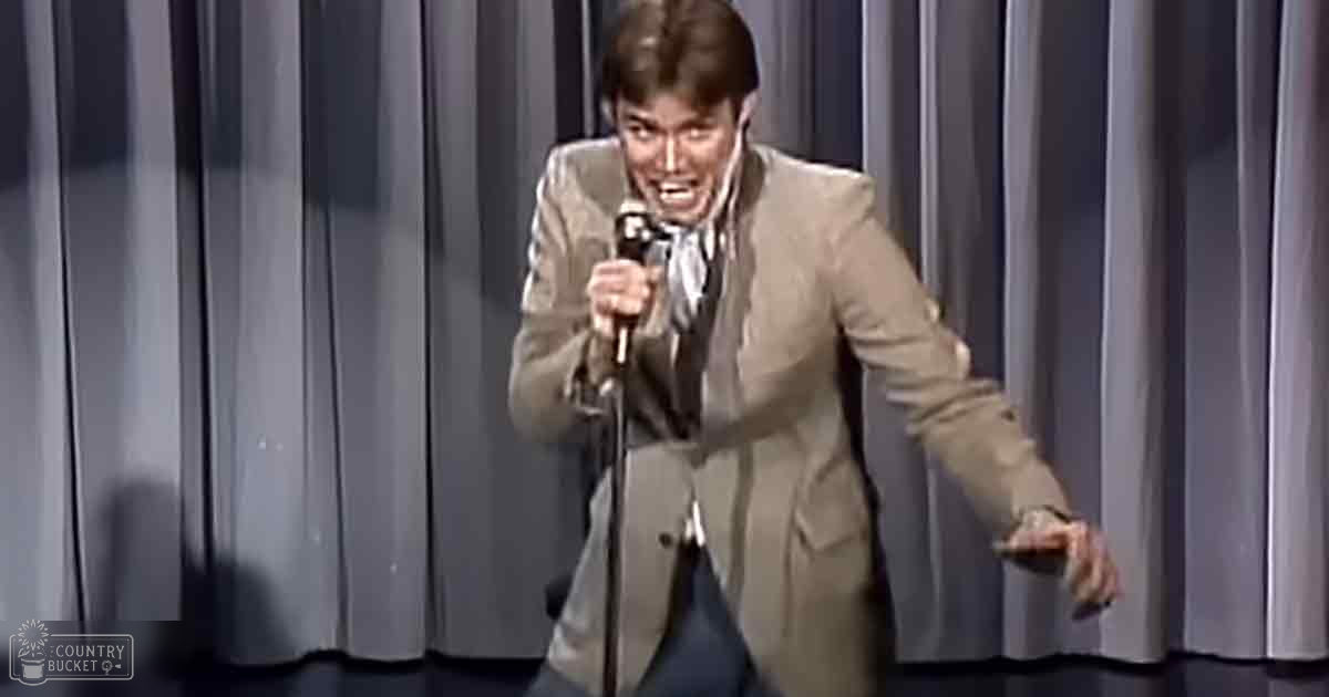 An adult male comedian in a beige suit performing stand-up comedy on stage with a plain curtain backdrop, suggesting a mid-20th century setting.