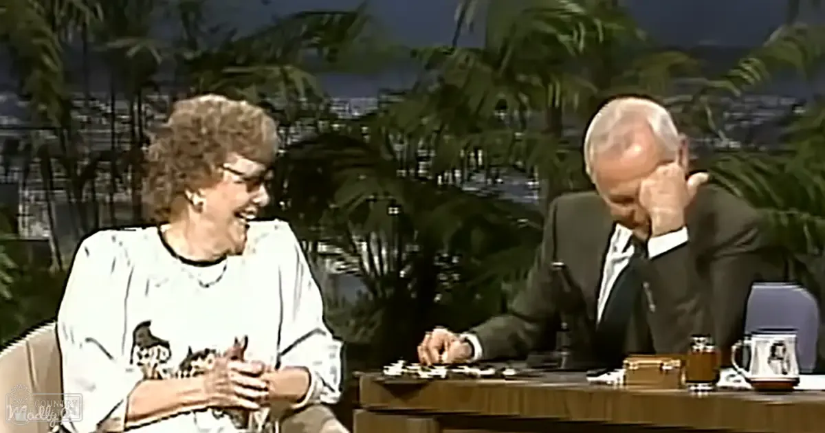 Two older adults seated at a desk with palm trees and cityscape backdrop, suggesting an outdoor studio setting with artificial lighting.