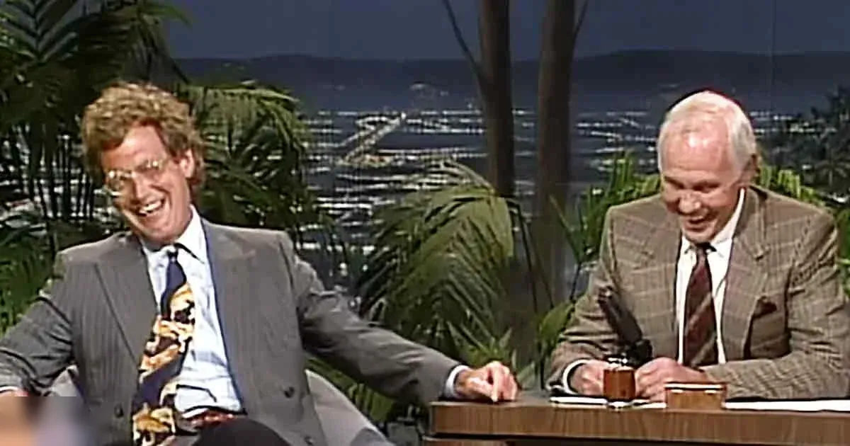 An adult male talk show host wearing a gray suit jacket over a light blue patterned tie with abstract shapes seated at a desk in a television studio, with another older adult male guest in a plaid suit and striped tie. Decorative plants and scenic city lights suggest an indoor set designed to mimic nature.