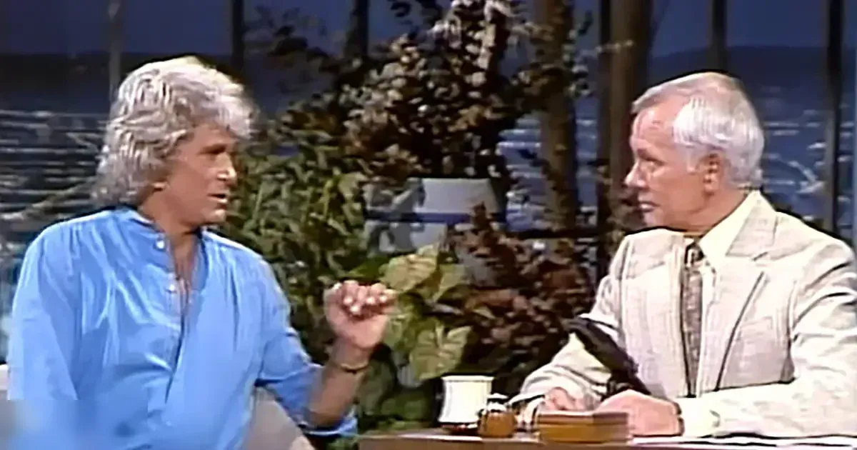A talk show host in a studio setting with another older adult male, both wearing formal attire; desk with mug and papers suggests an indoor professional environment.