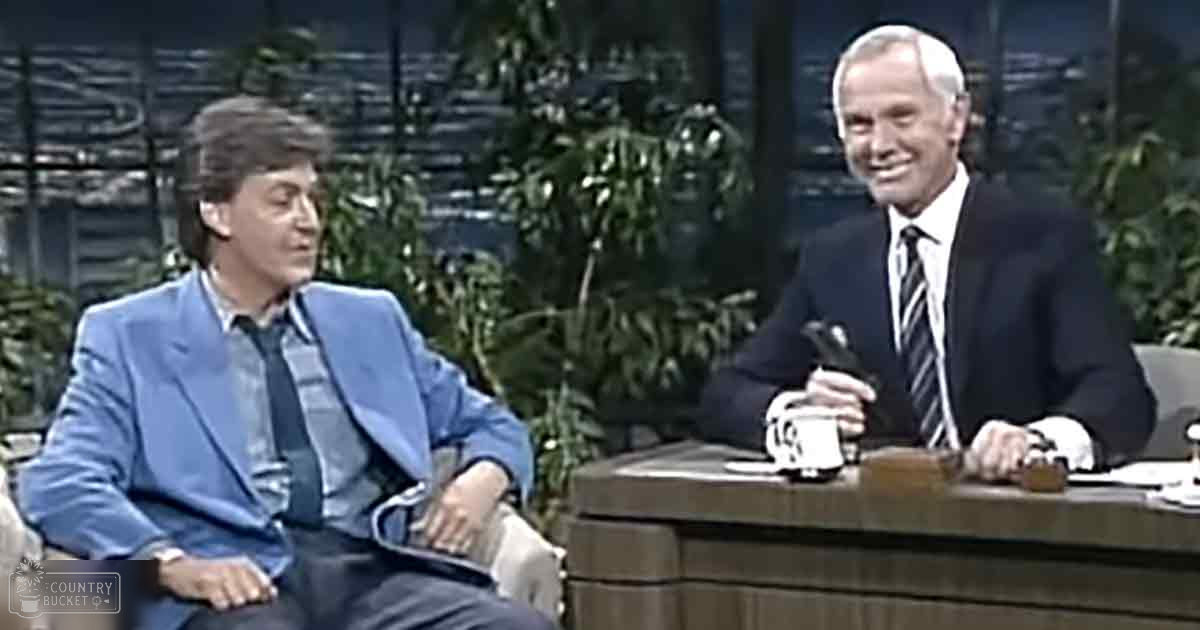 An adult male talk show host in a light blue suit jacket and an older male guest wearing a dark suit with a striped tie, seated at a wooden desk indoors.