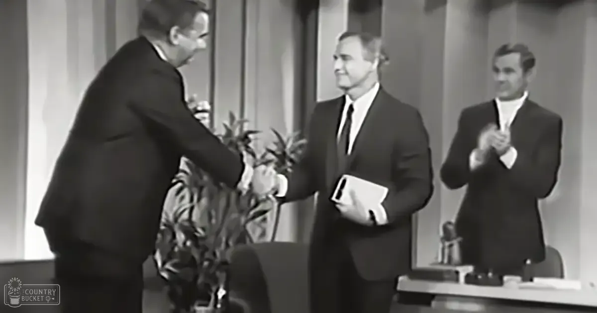 Indoor setting at what appears to be an awards ceremony or talk show; adults in dark suits, classic fit, and collared shirts with solid colors, possibly wool fabric, gathered around a stage setup with a microphone stand.