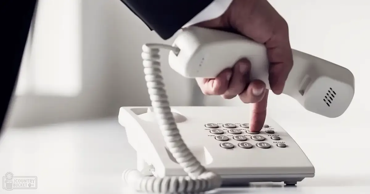 An adult male hand wearing a dark sleeve dials a standard office-style telephone indoors, with minimal decor in a plain light-colored setting.