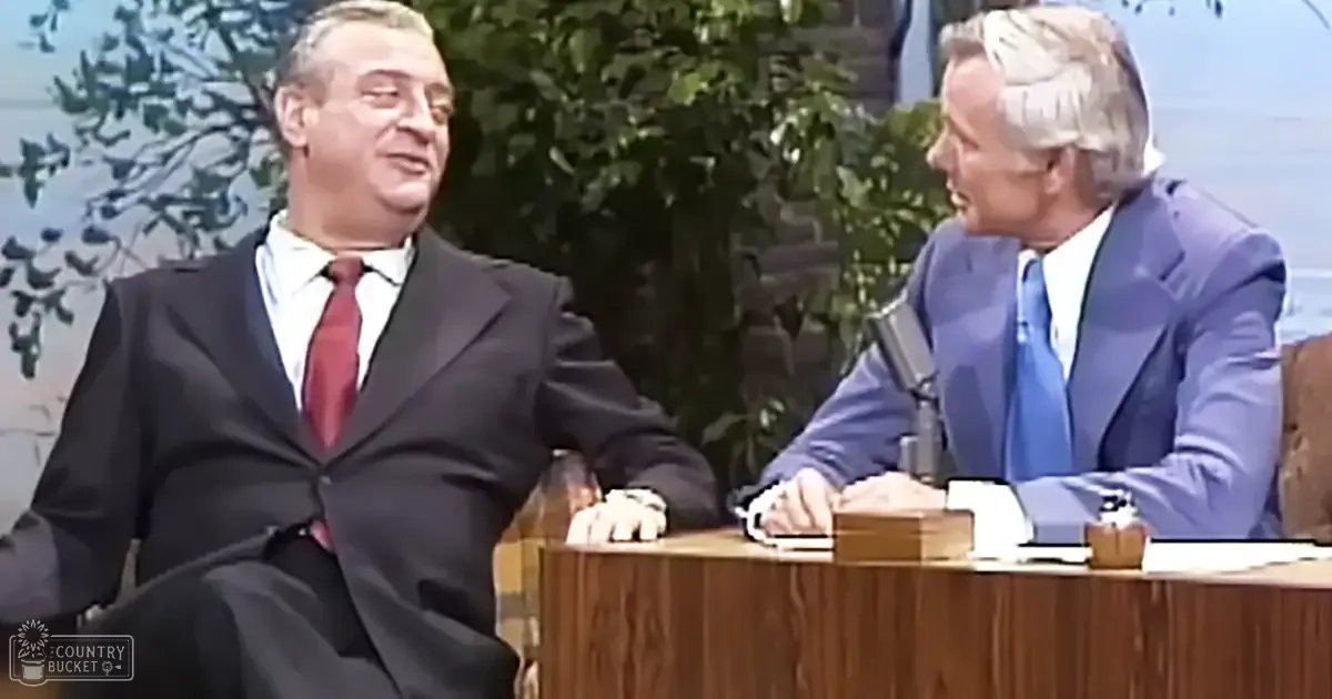 Two middle-aged to older adult men in a daytime indoor studio setting with a backdrop of trees and sky, wearing formal suits; one is seated at a desk with a microphone nearby.