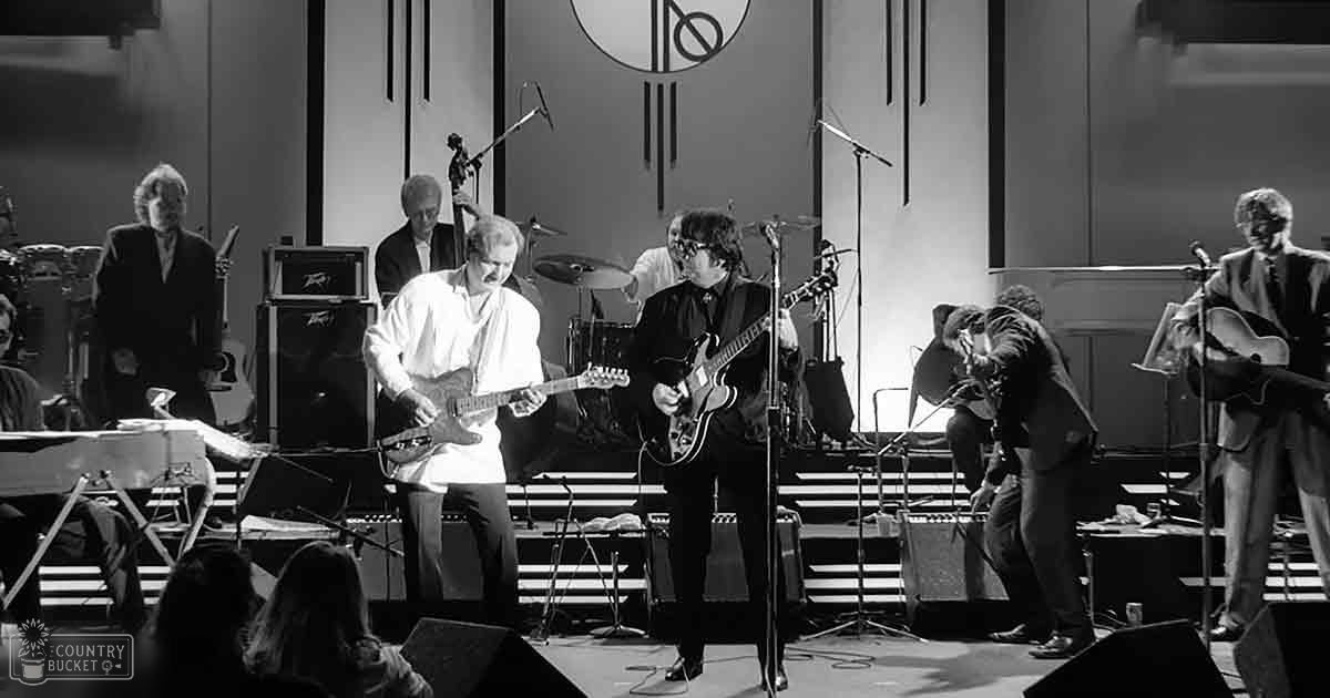 Musical performance in an indoor concert setting with performers wearing dark suits and playing electric guitars.