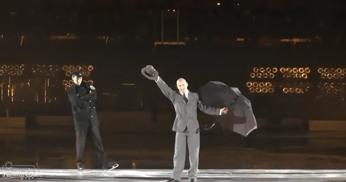 A performance scene at an indoor ice rink with two adult male performers dressed formally, one holding a microphone and the other an umbrella. The setting is illuminated by stage lighting against a dark backdrop.