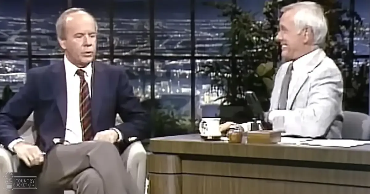 Middle-aged to older adult male in a dark suit jacket and patterned tie interviewing an older adult male in a light blazer over a white collared shirt, set against a backdrop of illuminated cityscape at night with greenery near the window.