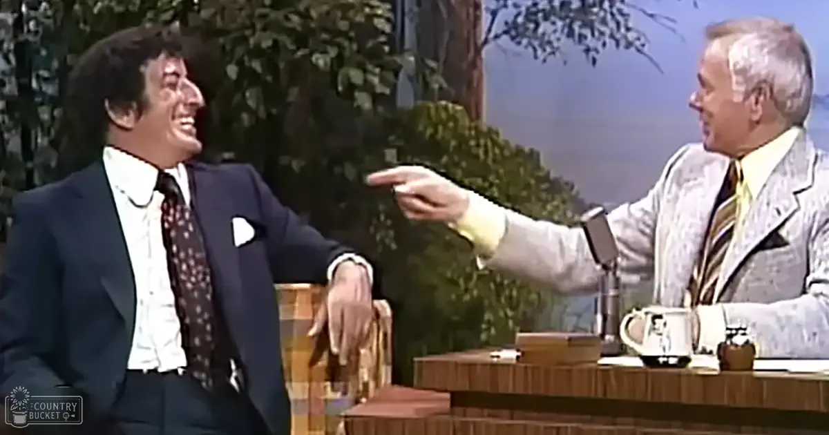 A middle-aged male talk show host interacts with an adult male guest outdoors, surrounded by greenery and a clear sky.