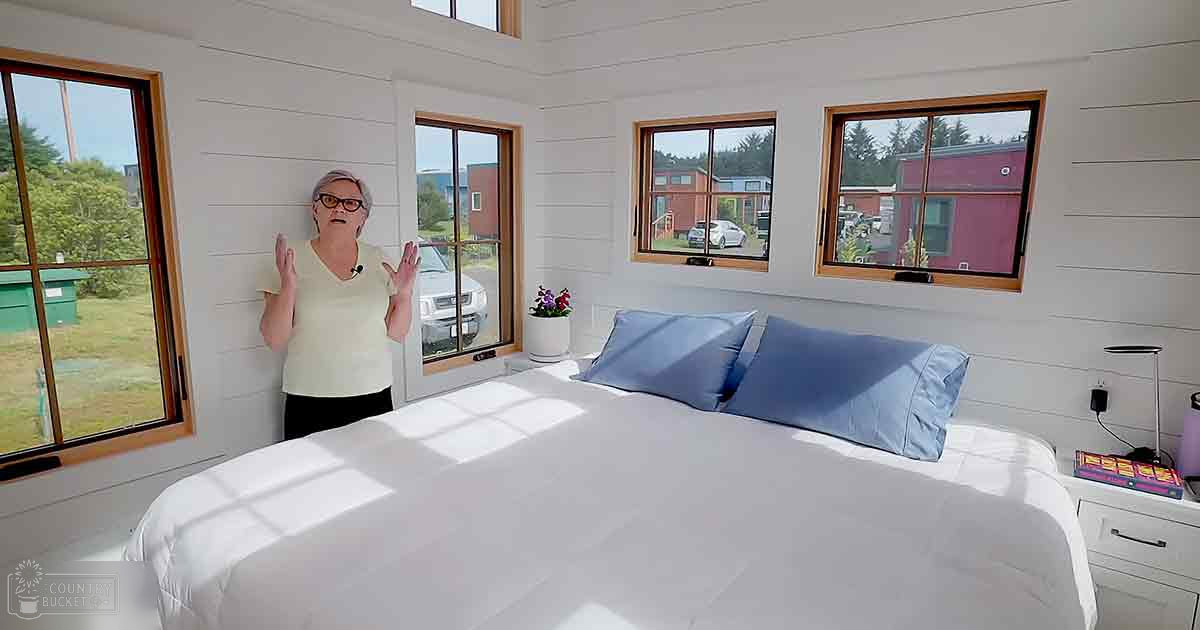 A middle-aged adult female in a bedroom setting with light yellow V-neck top and glasses, surrounded by white bedding, blue pillows, wooden windows, and outdoor greenery. The natural daylight suggests daytime during spring or summer.