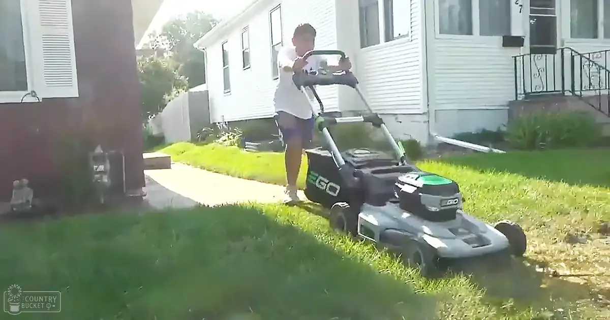 A young adult male mowing grass in a suburban neighborhood during late afternoon or early evening, wearing a white t-shirt and dark shorts.