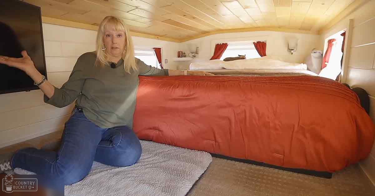 A middle-aged woman in a cabin, wearing a long-sleeved green shirt and blue jeans, kneeling near a bed with an orange blanket. The setting is rustic with wooden walls and ceiling, natural daylight streaming through red-curtained windows.