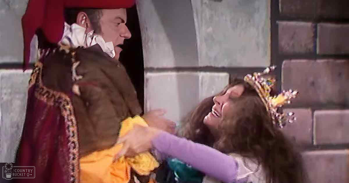 A child in a medieval setting wearing a purple sweater and a crown, accompanied by an older adult male in a brown tunic with red hat and fur-lined cloak, surrounded by stone walls suggesting a castle or fortress interior under bright daylight.