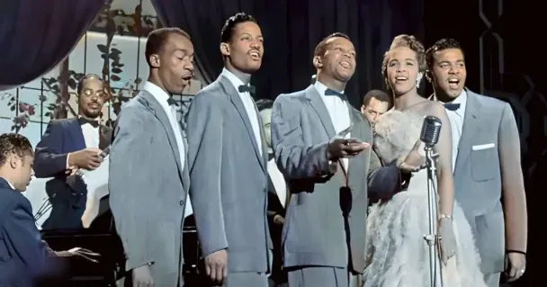 A lively musical performance scene indoors at night, with young adults in formal attire on stage with vintage-style microphone and piano visible.