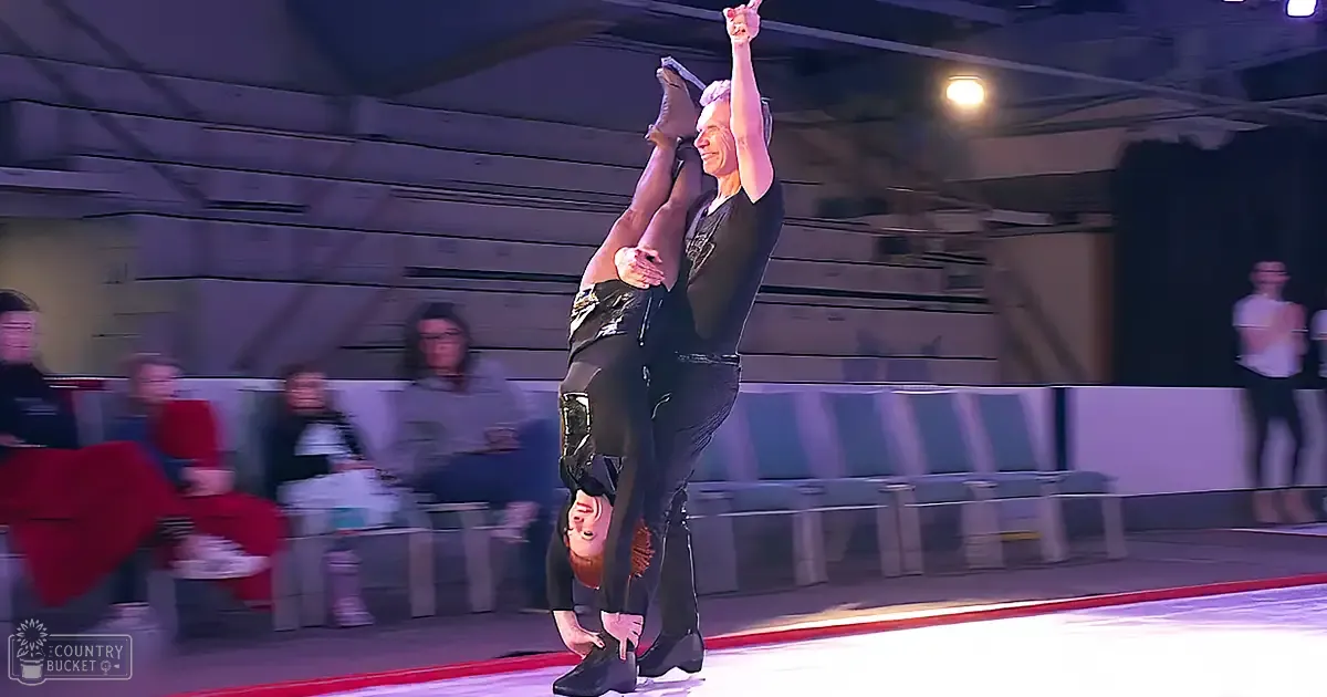 Male skater performing a dynamic move with his female partner upside down on an indoor ice rink filled with spectators under bright lighting.