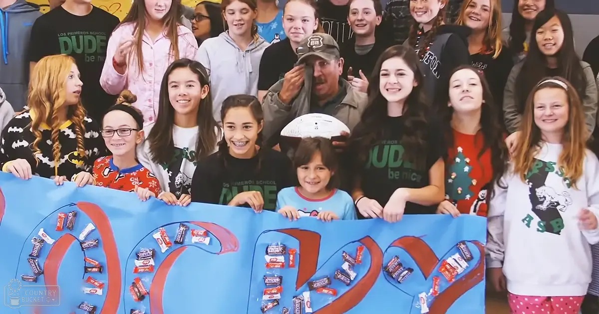 A group of teenagers at Los Primeros School, wearing festive shirts and holding white frisbees; a large blue banner with candy bars spelling '100' is prominent in the background.