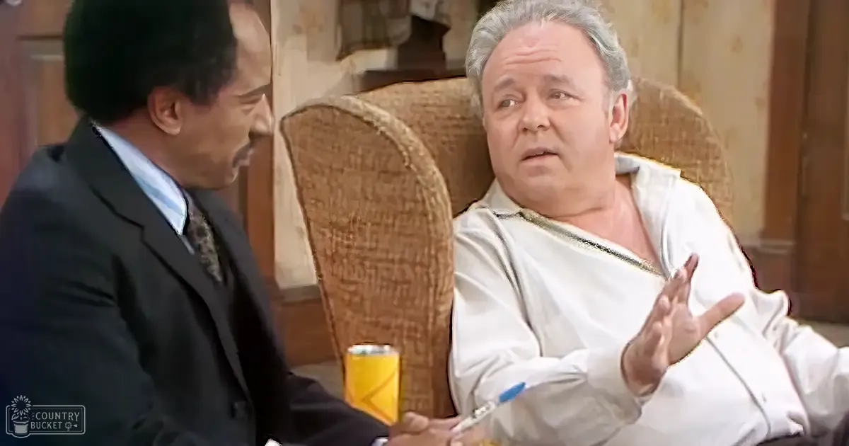 A middle-aged adult male in a suit stands beside an older man wearing a patterned shirt, seated on a brown armchair with a yellow canister nearby. Warm indoor lighting suggests a cozy living room setting.