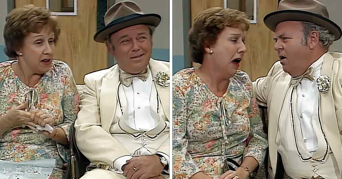 A middle-aged man in a formal white suit with decorative buttons sits next to an older adult woman wearing a floral-patterned blouse indoors near a wooden cabinet and framed artwork.