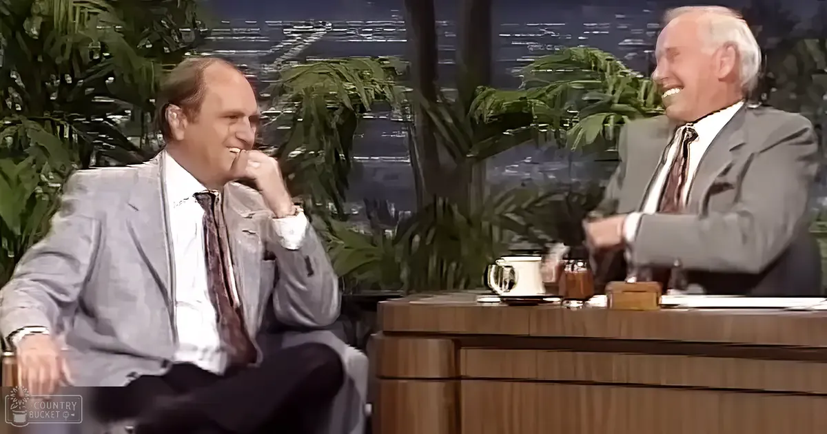 A middle-aged man wearing a light gray suit jacket sits on stage with an older adult male guest in a dark suit, both in a professional television studio set up for a talk show. The setting includes wooden furniture and large green plants.