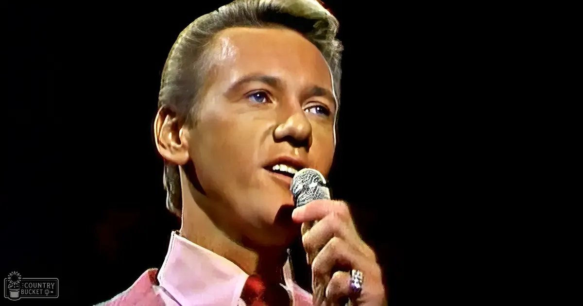 A middle-aged male performer in a light pink dress shirt and red tie, holding a microphone with a diamond-like pattern on stage at night.