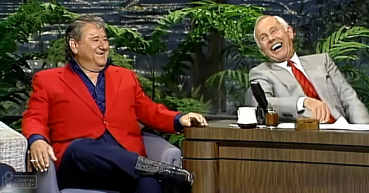 A middle-aged man wearing a bright red blazer over a dark blue shirt and pants sits on a couch, while an older adult male in a gray suit with a white shirt and red tie is seated behind a wooden desk. The setting appears to be indoors, possibly in a studio.