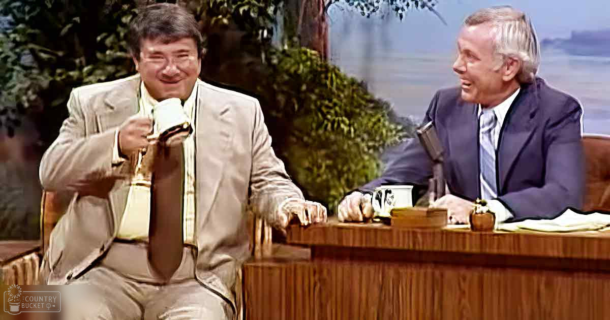 Talk show host and guest in a studio with wooden desk, microphones, and plants suggesting an outdoor theme.