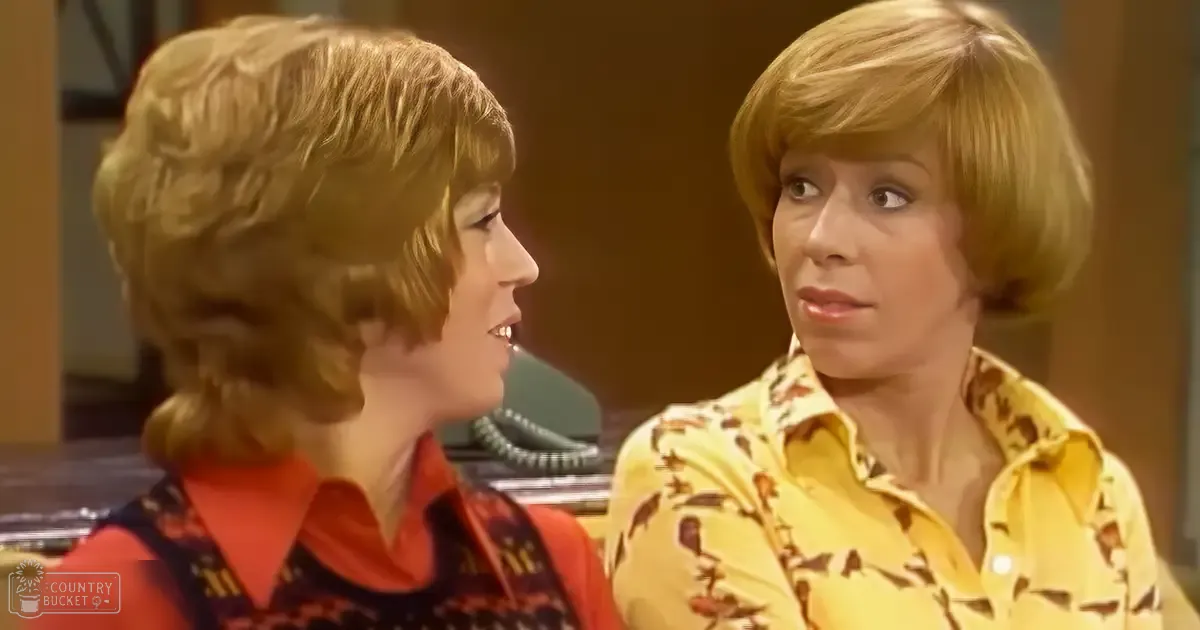 Two middle-aged women wearing loose-fitting 1970s-style blouses, seated indoors with a patterned sofa and vintage telephone, suggesting a domestic living room scene.