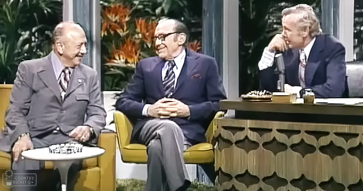 Three men seated in what appears to be a talk show set from the 1960s or 1970s, with modern yellow armchairs and tropical plants in the background.