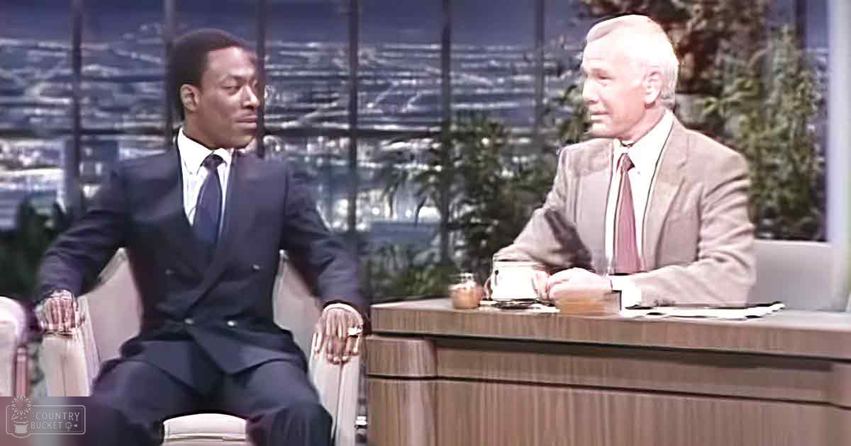A television studio setting with two male adults, one wearing a dark blue suit and another in a beige suit jacket over white collared shirts; cityscape backdrop at night.
