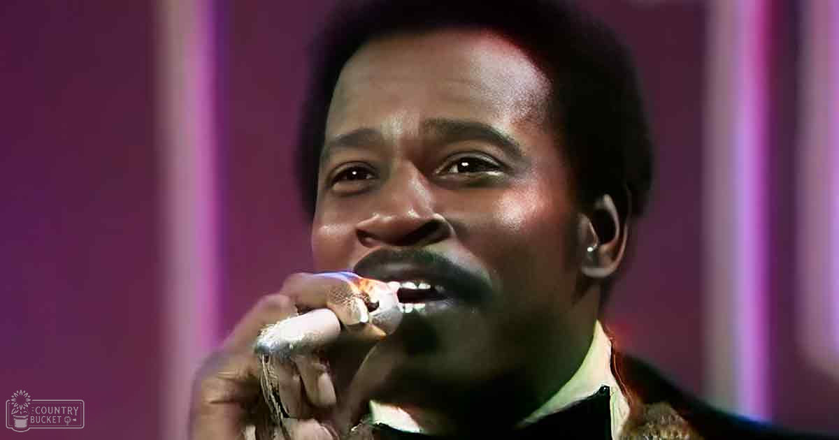 An adult male singer in a dark, patterned, textured garment from the 1970s or early 1980s holds a white-covered microphone on stage with vertical purple stripes in the background.