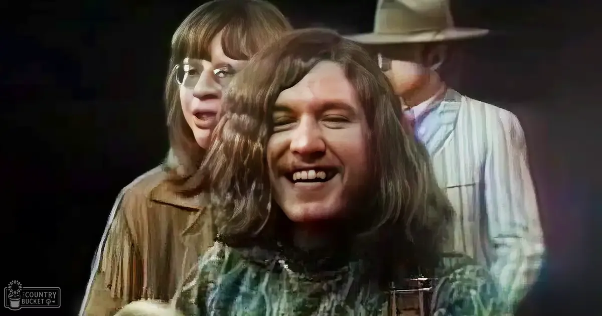 A young adult male with long brown hair and a greenish plaid shirt smiles indoors while wearing glasses. A child or teenager, possibly female, stands in the background wearing a brown jacket with fringes.