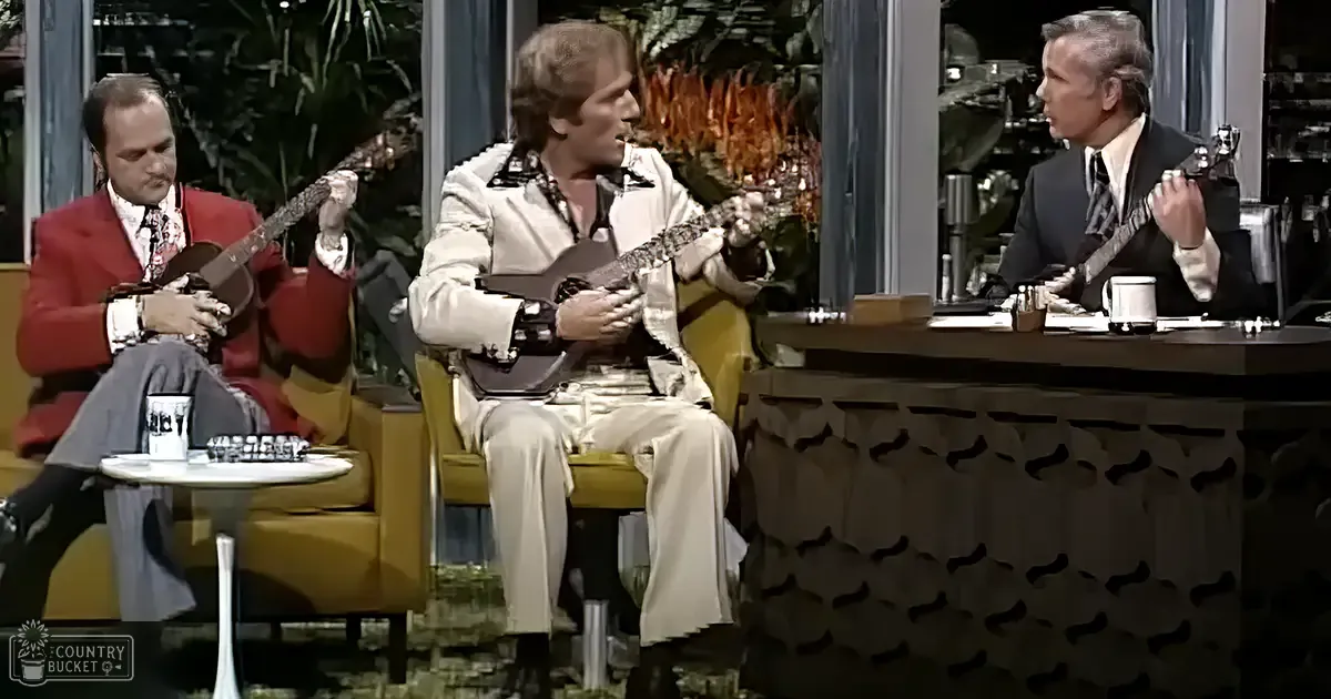 An indoor studio with yellow armchairs, a table holding a mug and small items, plants visible through large windows, and musical instruments being played by two adults in formal attire.
