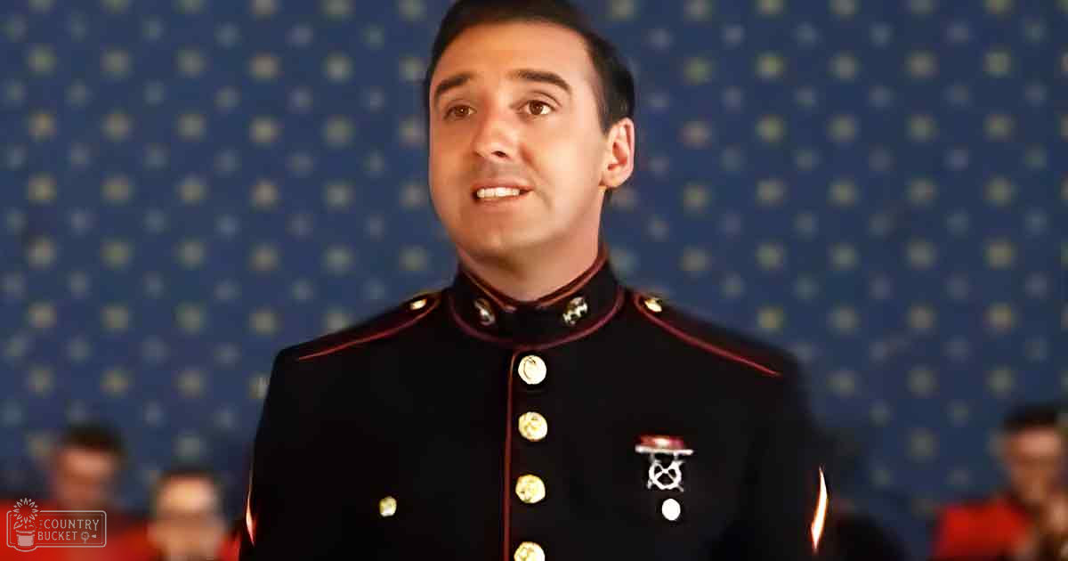 Young adult male wearing a dark blue military-style dress uniform with red trim, gold buttons, epaulets, and an insignia near the chest at an indoor event or performance setting.