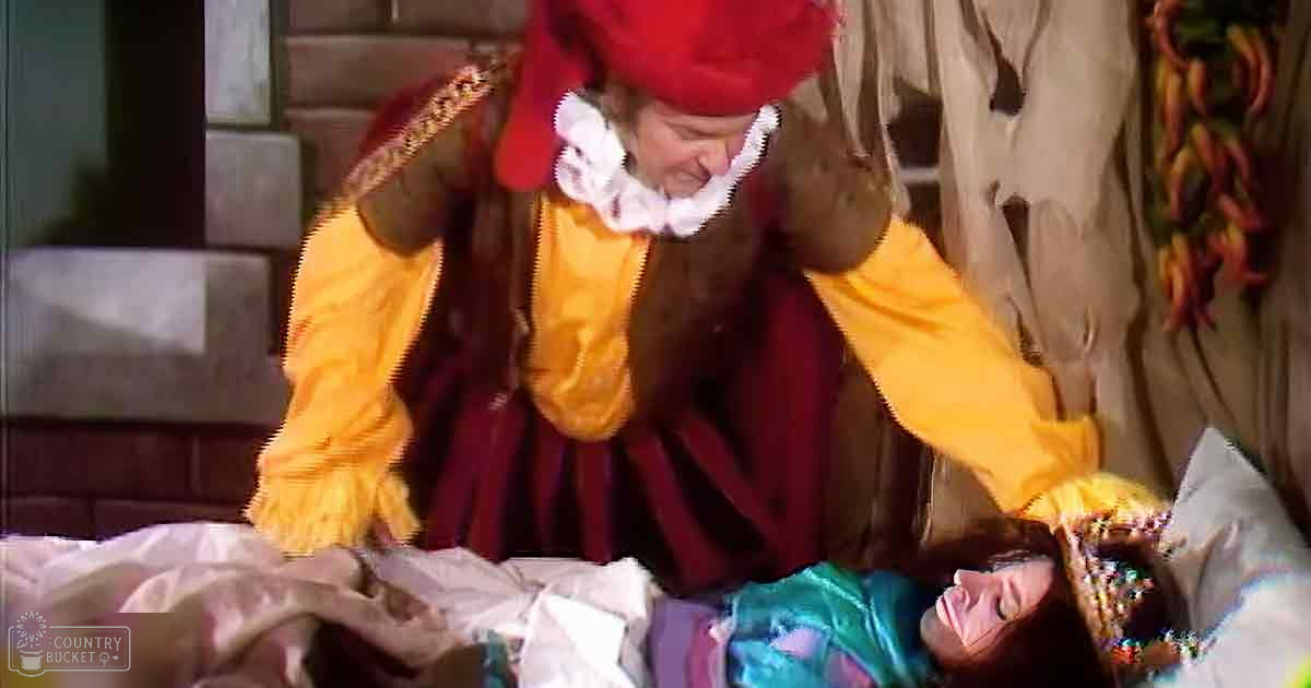 A middle-aged male clown in a theatrical setting with an oversized yellow and red clown costume, interacting with a young adult female in a brightly colored striped dress. The scene is indoors on a rustic stage with brick walls and dried chili peppers.