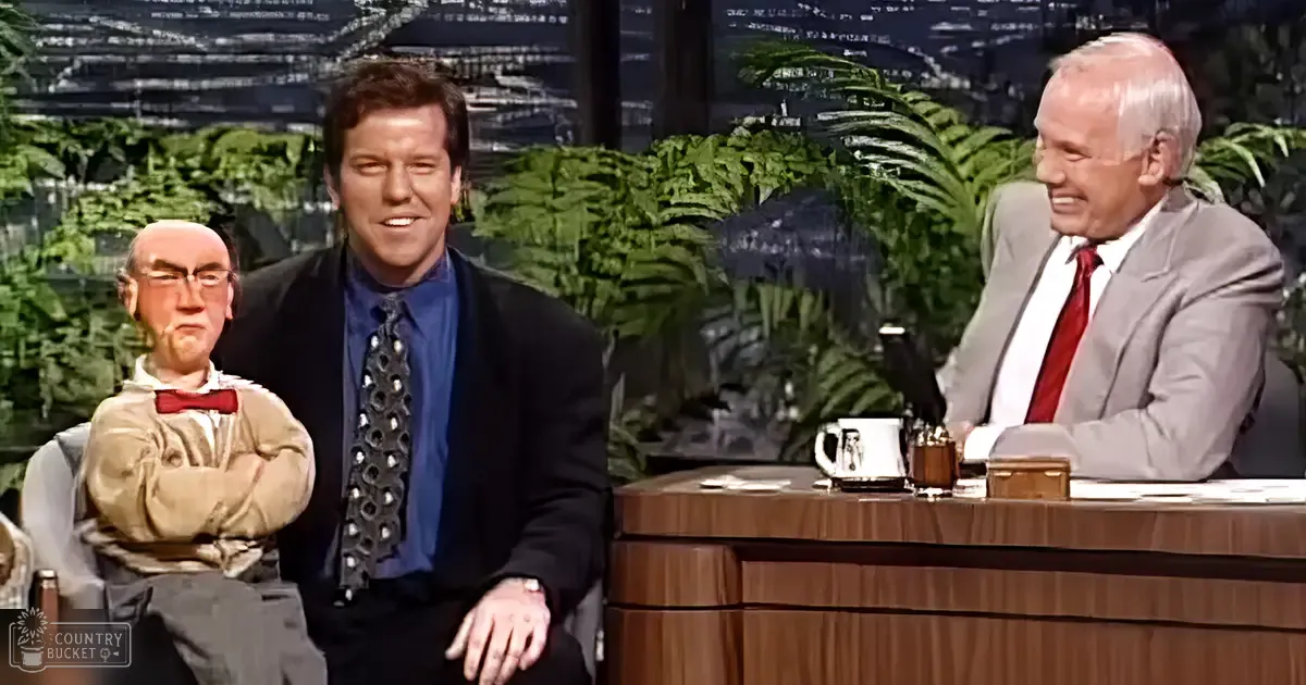 Talk show host in an indoor studio with greenery and cityscape visible through windows, with a wooden desk and papers, set against professional stage lighting.