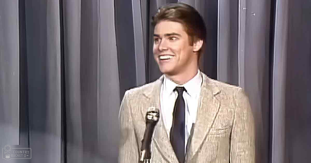 A young adult male in a mid-20th-century formal suit stands on an indoor stage with a microphone, under even and bright lighting.