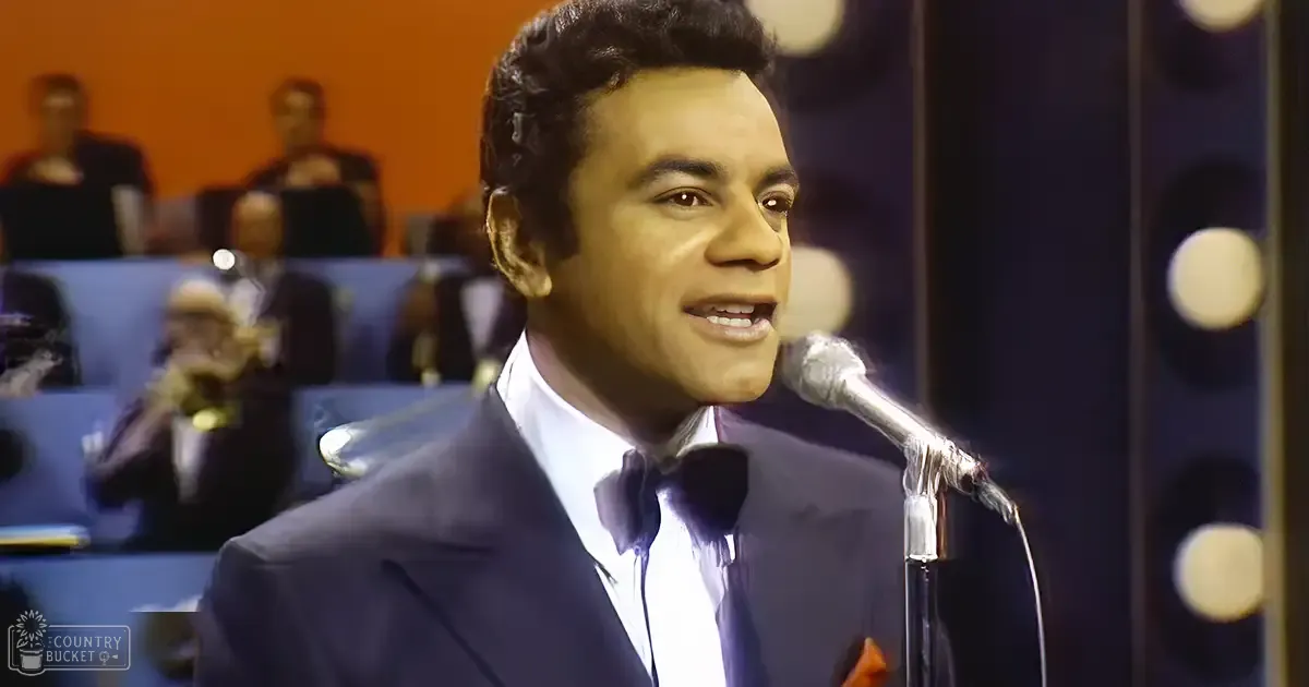 A male adult singer wearing a dark suit with white shirt and bow tie, singing into a microphone in what appears to be a professional recording or performance studio environment.