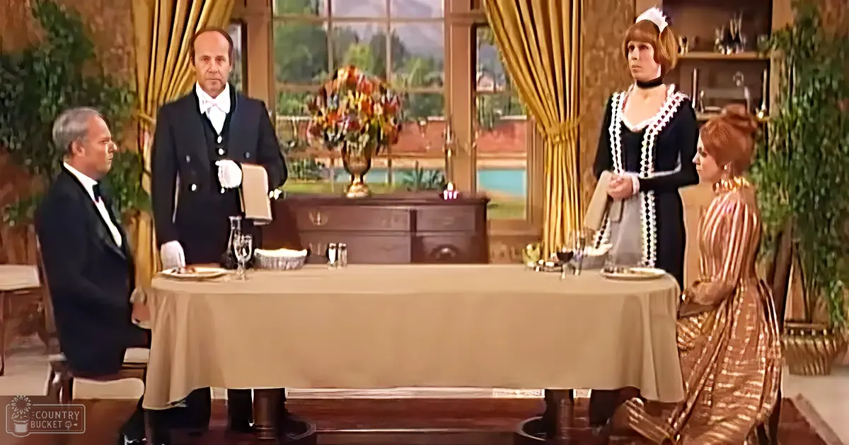 A butler in a dark tuxedo and an adult female maid uniform are seated at a table in what appears to be a dining room or lounge area, with another adult male standing nearby holding a menu.