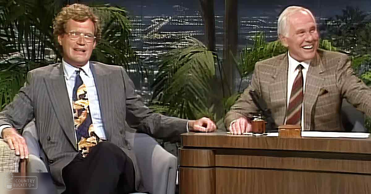 Adult male talk show host and guest in a studio setting, wearing light gray pinstripe suit over white shirt with colorful animal print tie; older adult male seated behind desk in brown plaid blazer, white collared shirt, red striped tie. Indoor studio environment with greenery and cityscape lighting.
