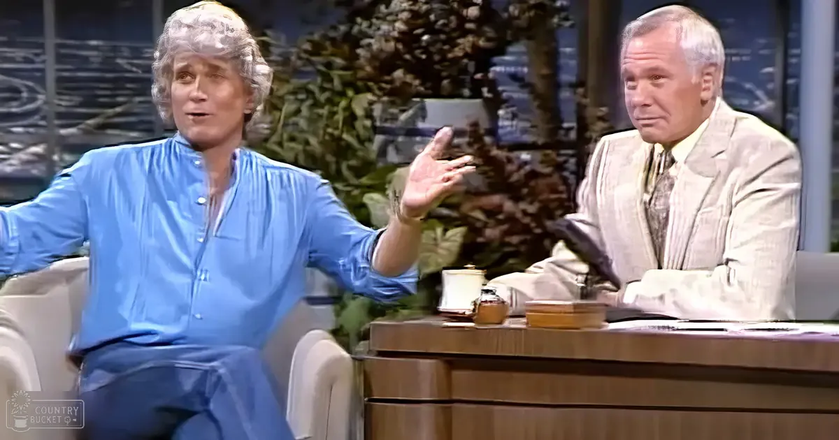 Two adults in a television studio setting, one middle-aged man wearing casual attire with blue button-up shirt and dark pants, seated on a white armchair; another older adult male dressed formally in beige suit jacket over striped dress shirt and tie, seated at a wooden desk with professional lighting and decorative elements.