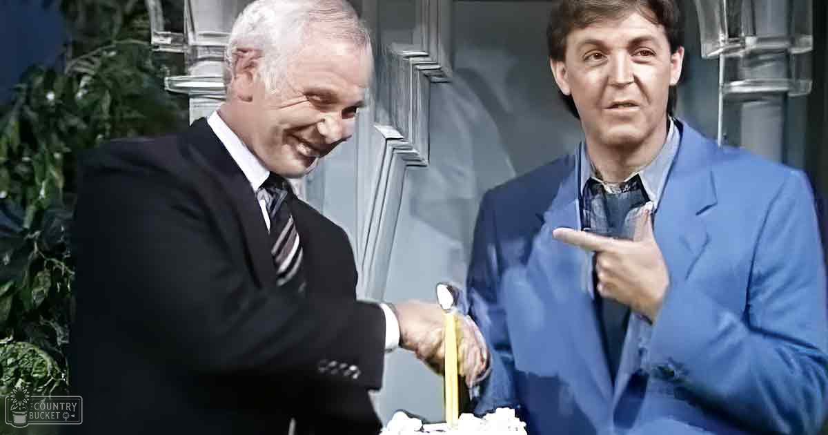 Two adult males dressed formally, one holding a small object near a lit candle on a cake at an indoor celebration with decorative plants.