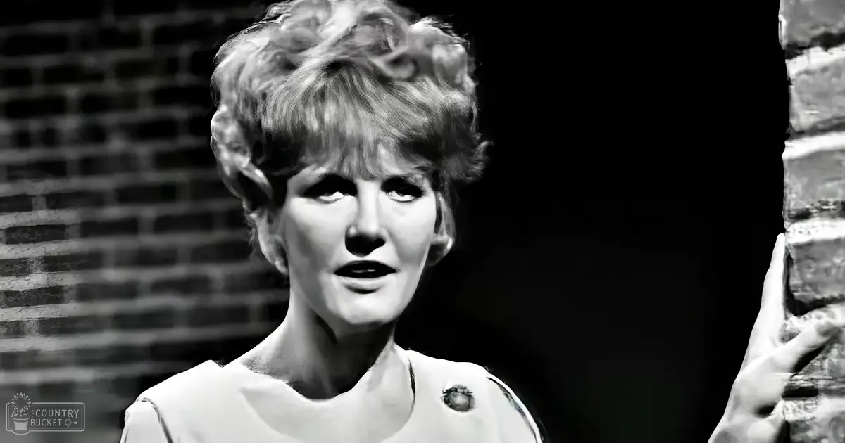 A middle-aged female wearing a white garment with short sleeves and a small dark circular detail on the left chest stands against a dark brick wall in an indoor setting, illuminated by dramatic lighting.